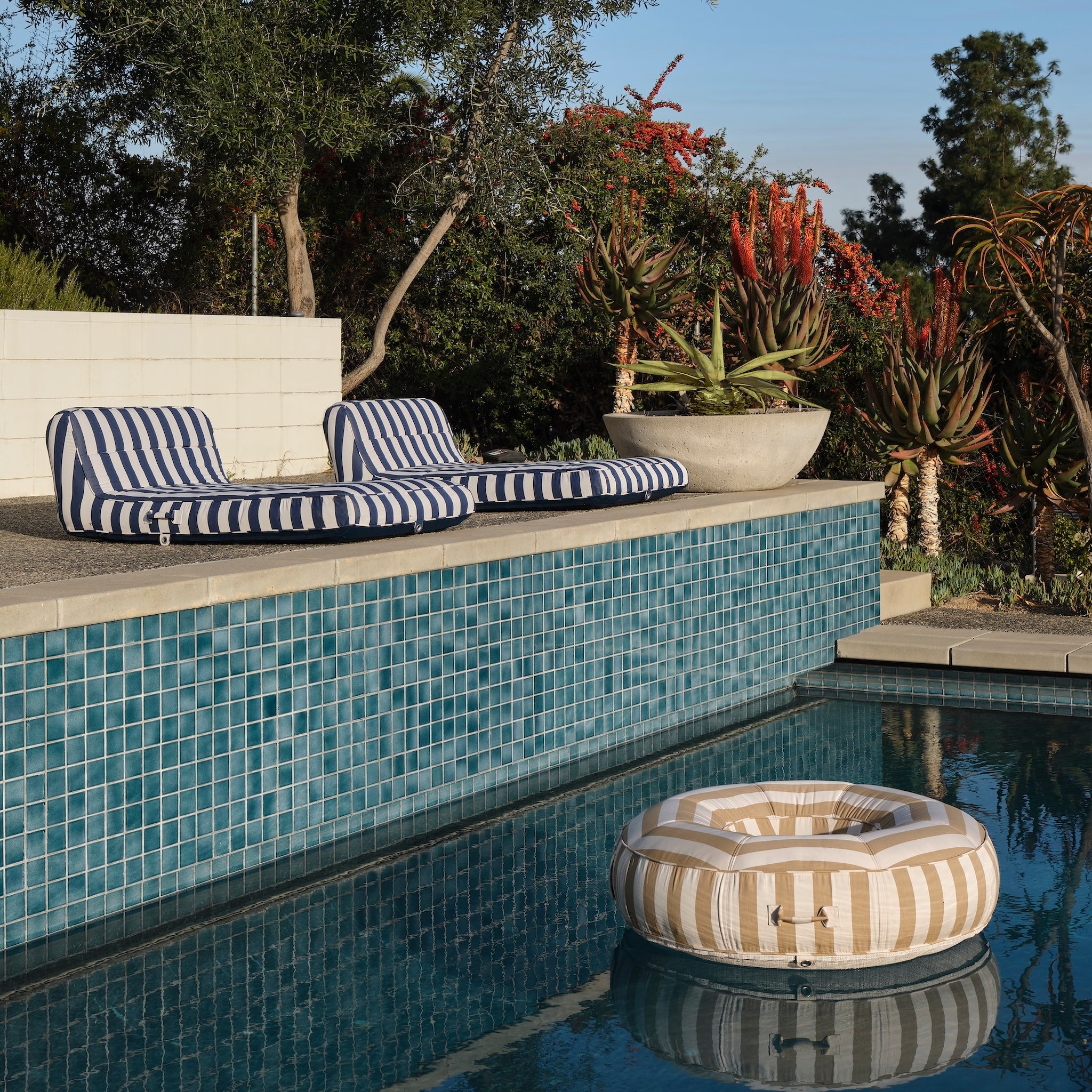 Two blue and white single luxury pool floats by a swimming pool with a beige and white luxury pool float floating in the swimming pool.