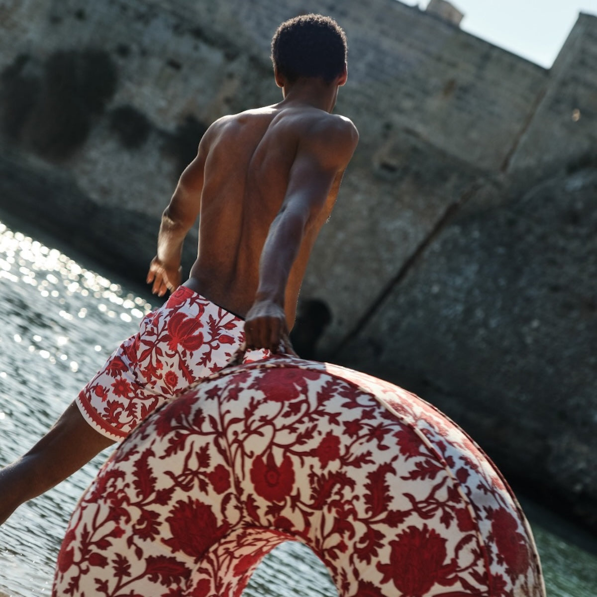A man swinging a luxury red and white pool float on a beach in Pulgia, Italy.