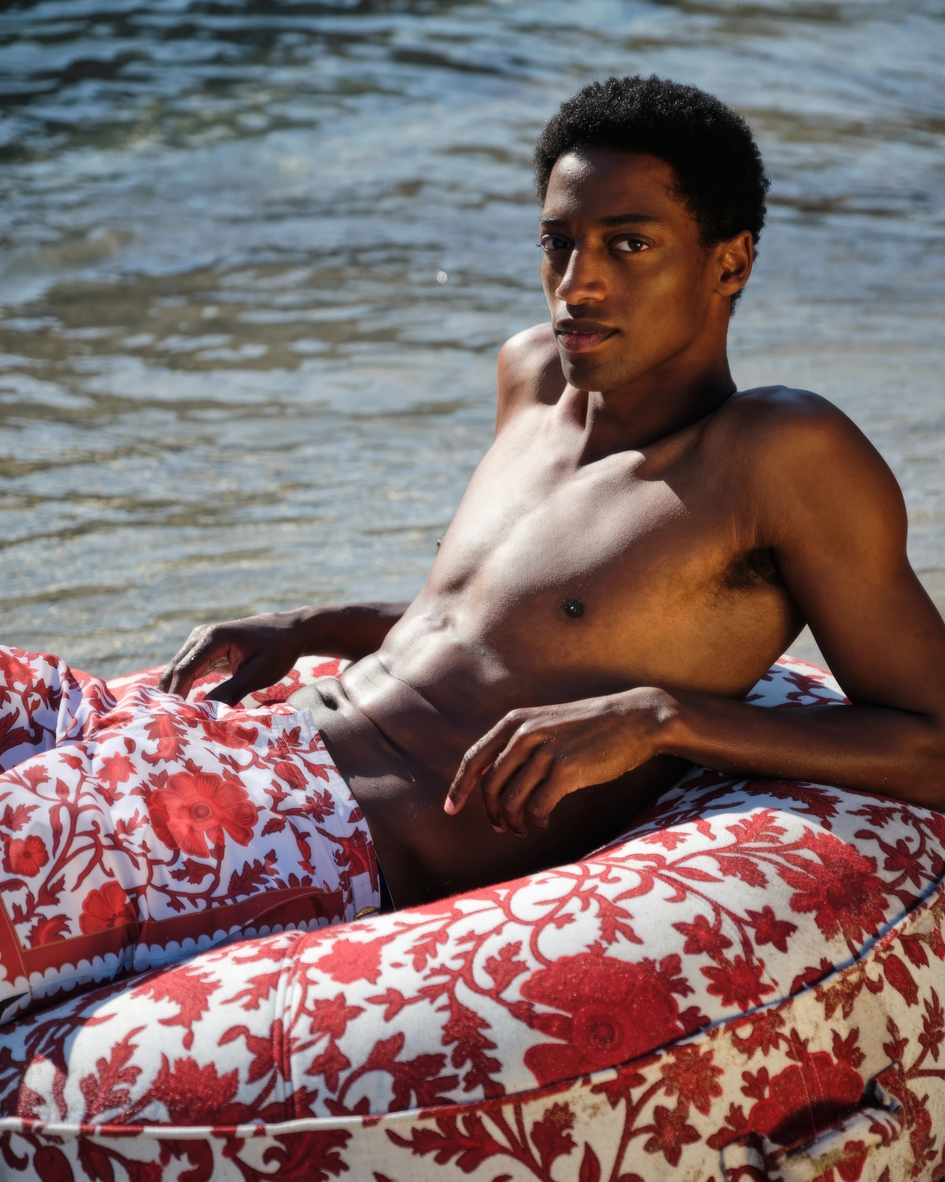 Model lounging confidently on a designer circular pool float with bold red floral upholstery from Oliver James Lilos, styled for elegant summer relaxation.