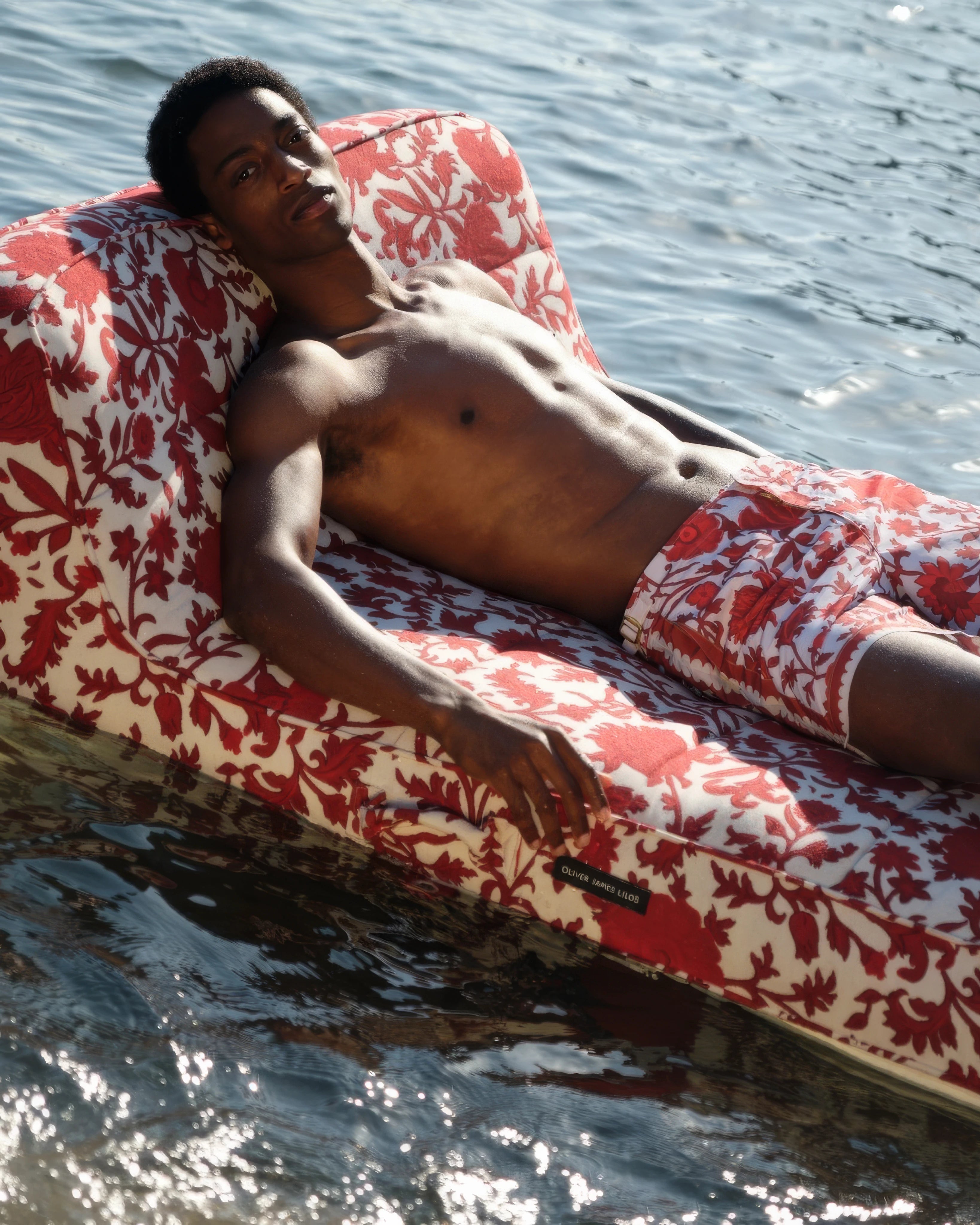 Man reclining on an Oliver James Lilos inflatable lounger in matching red and white baroque swimwear, drifting on the surface of a tranquil lake.