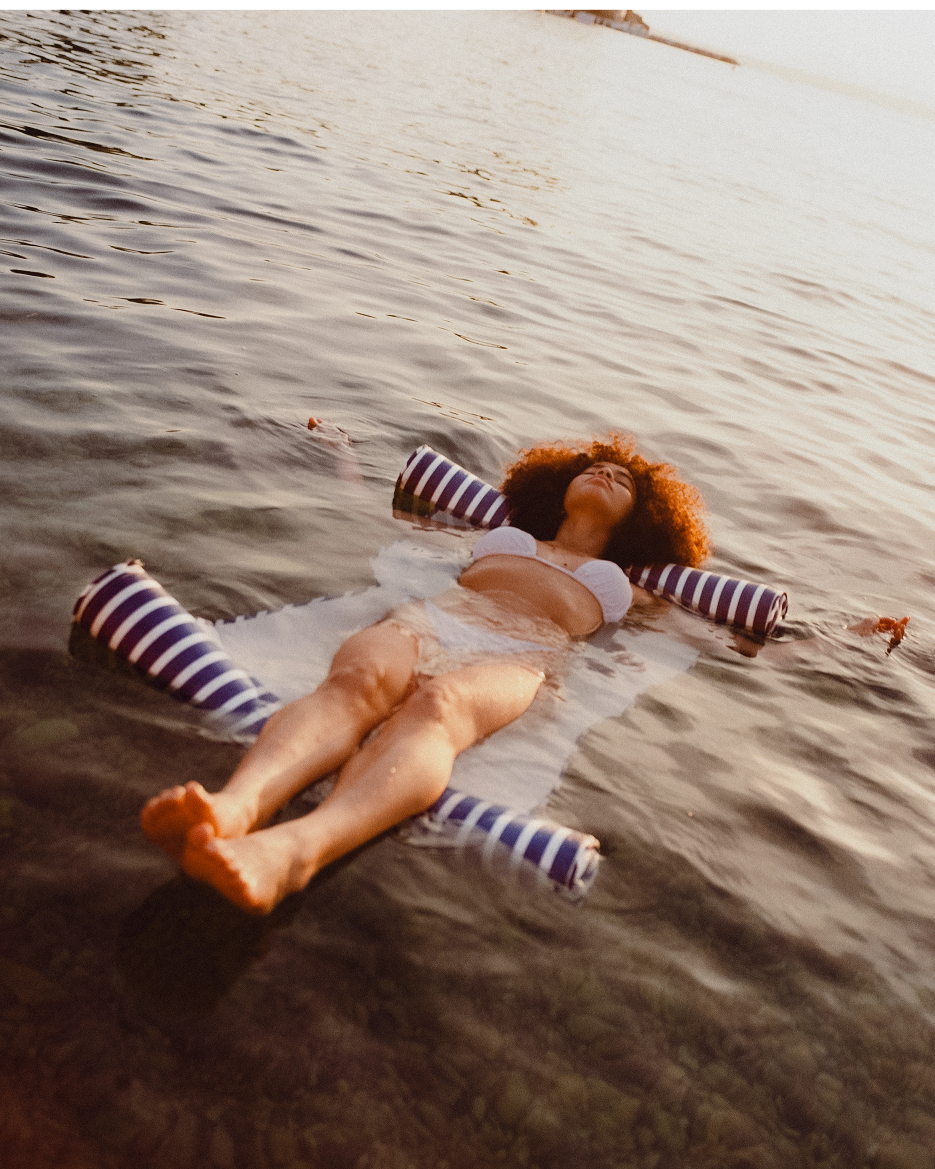 A woman with natural curls floats peacefully at sea on a striped floating hammock, wrapped in golden light — where coastal tranquility meets refined leisure