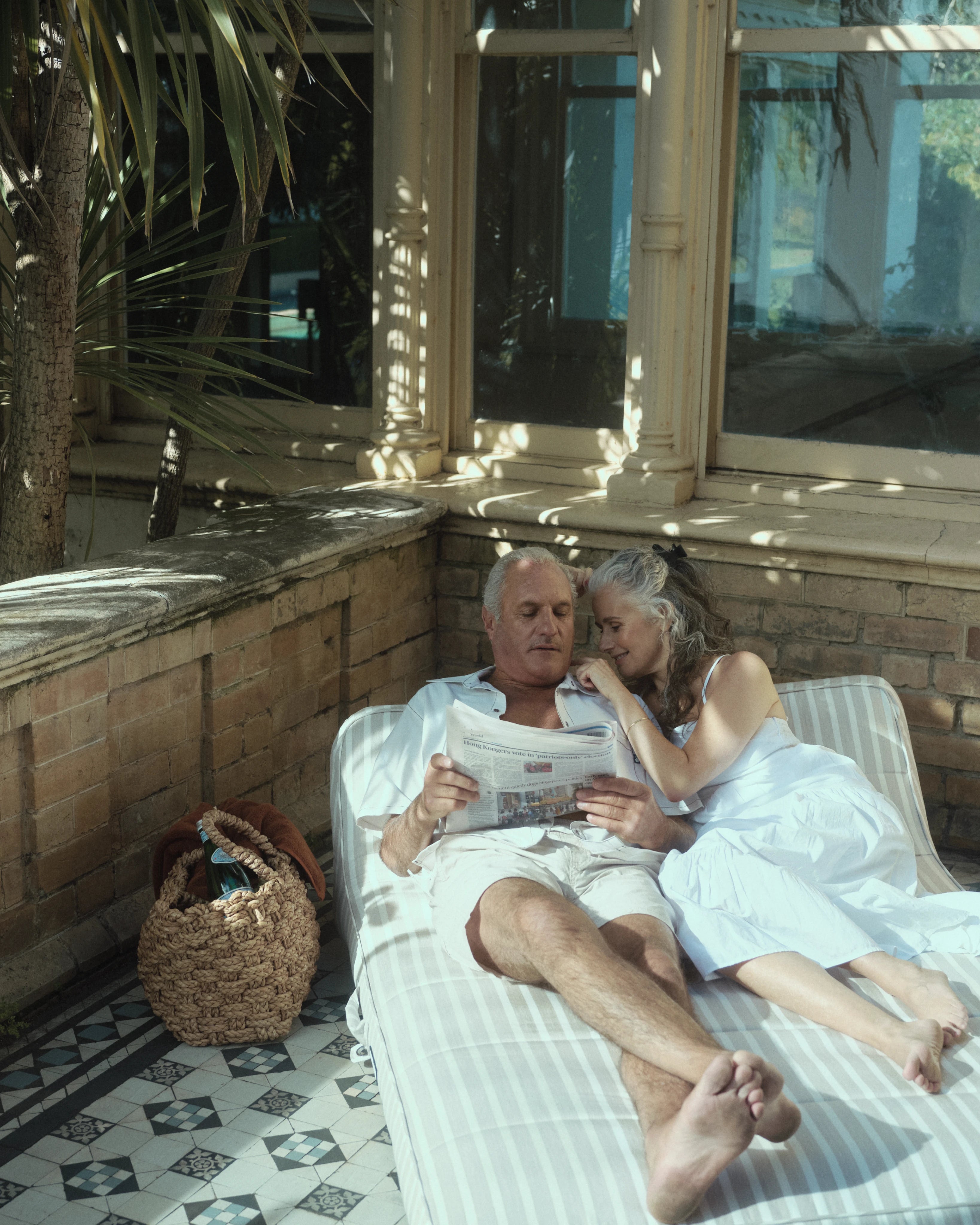An older couple lounges together on a double luxury pool float in a tiled courtyard, wrapped in soft sunlight and timeless affection — a scene of quiet intimacy and enduring style.