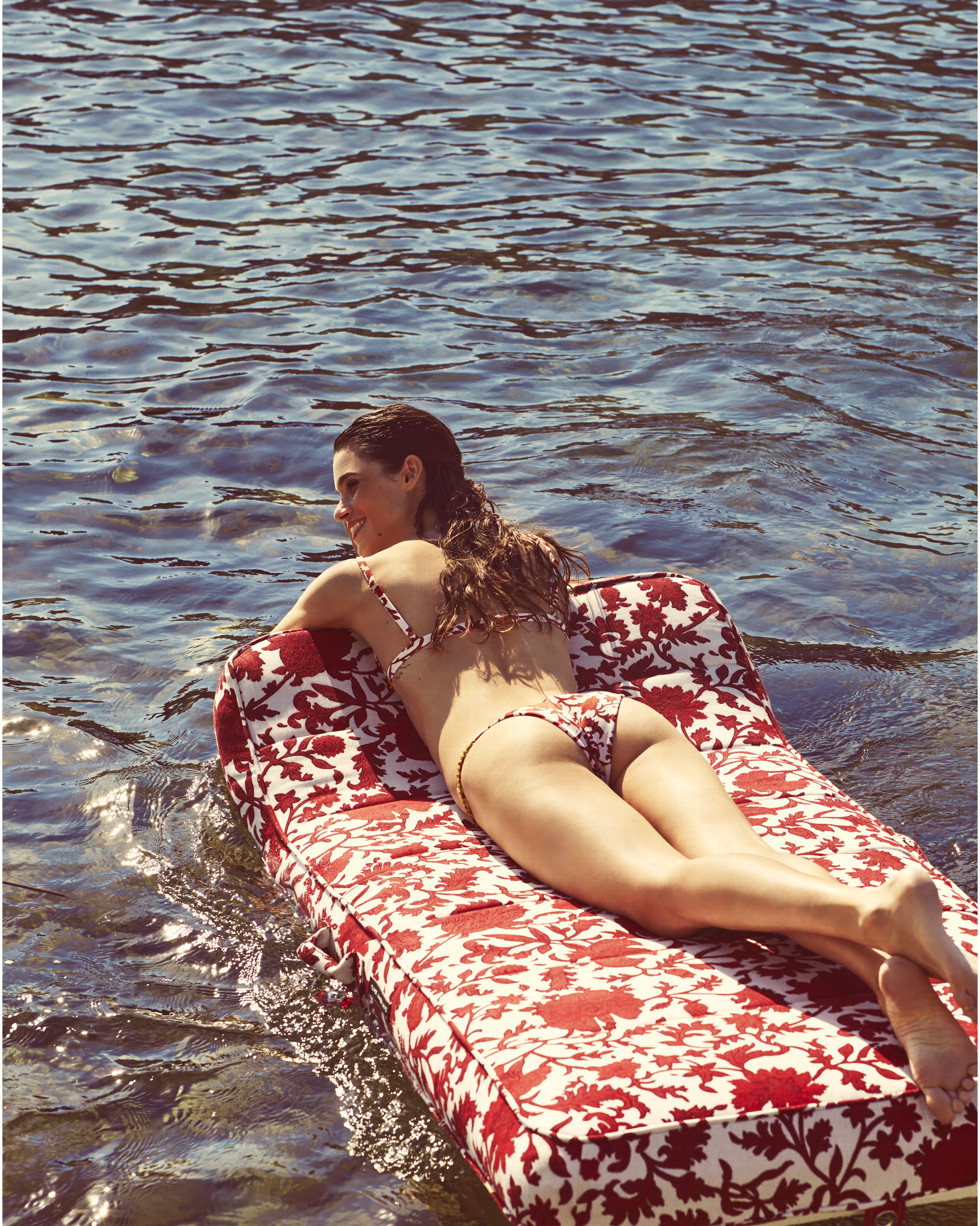 Woman sunbathing on a red and white damask-patterned luxury pool float by Oliver James Lilos, floating on a serene lake.