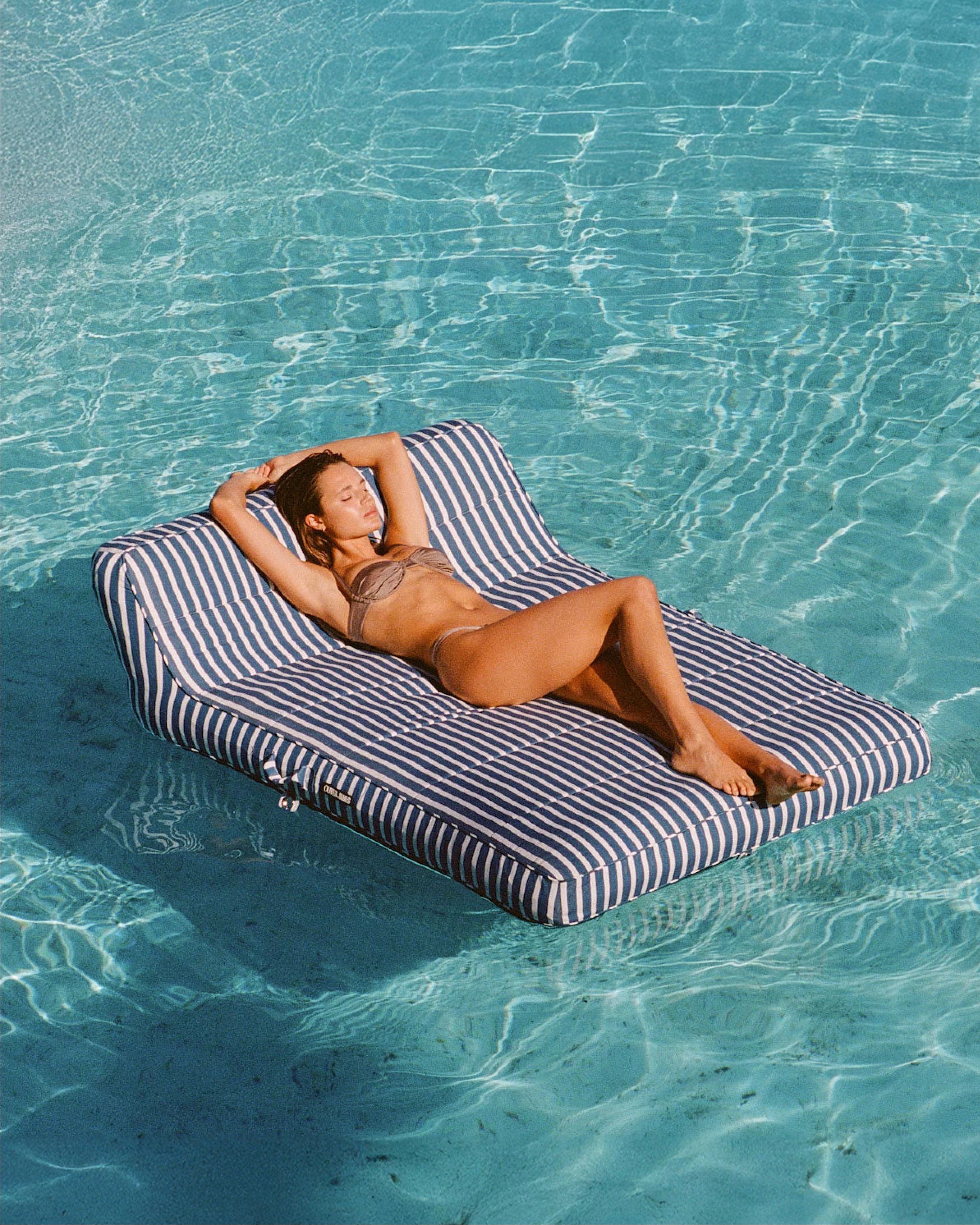 Bathed in sunlight, a woman reclines on a navy striped luxury pool float by Oliver James, surrounded by calm turquoise water — pure, poolside serenity.