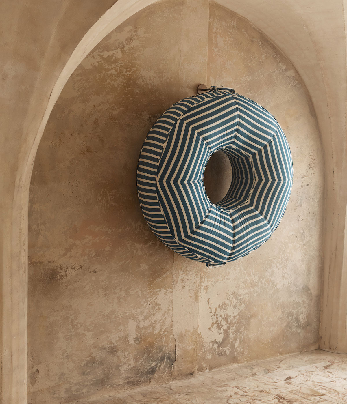 A luxury pool float ring lounger hanging beneath a marble stone archway by an eyelet made of teak and stainless steel.