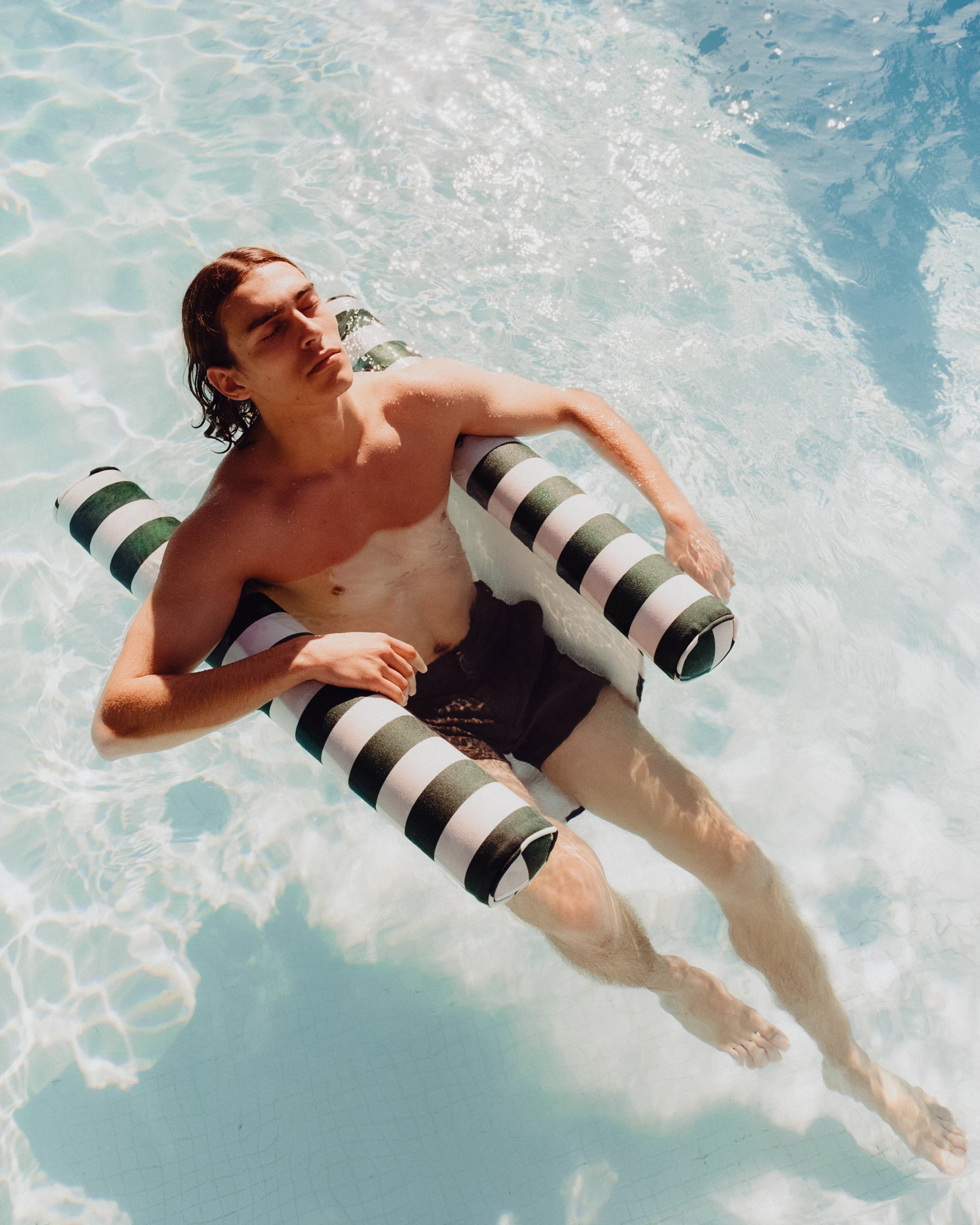 Man relaxing on a striped floating hammock in a sunlit pool — designer water lounger combining elegance, comfort, and effortless buoyancy.