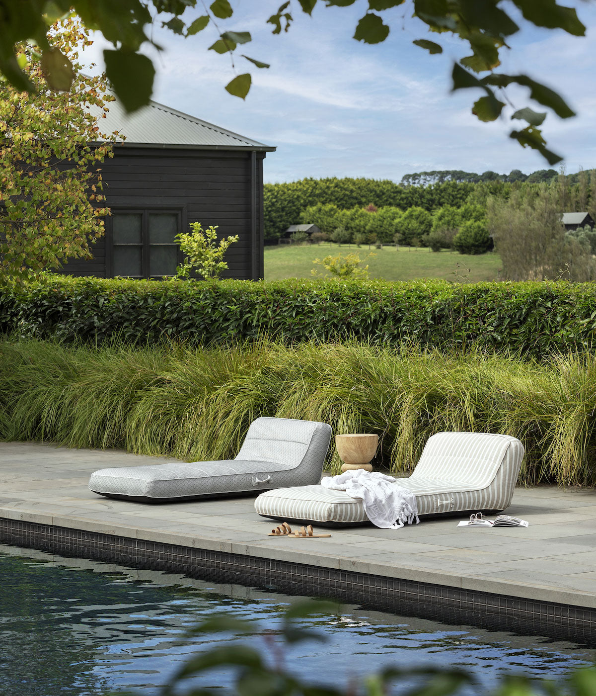 Two pool floats for adults sitting by a swimming pool, styled with a towel and slippers with rolling green hills in the background.