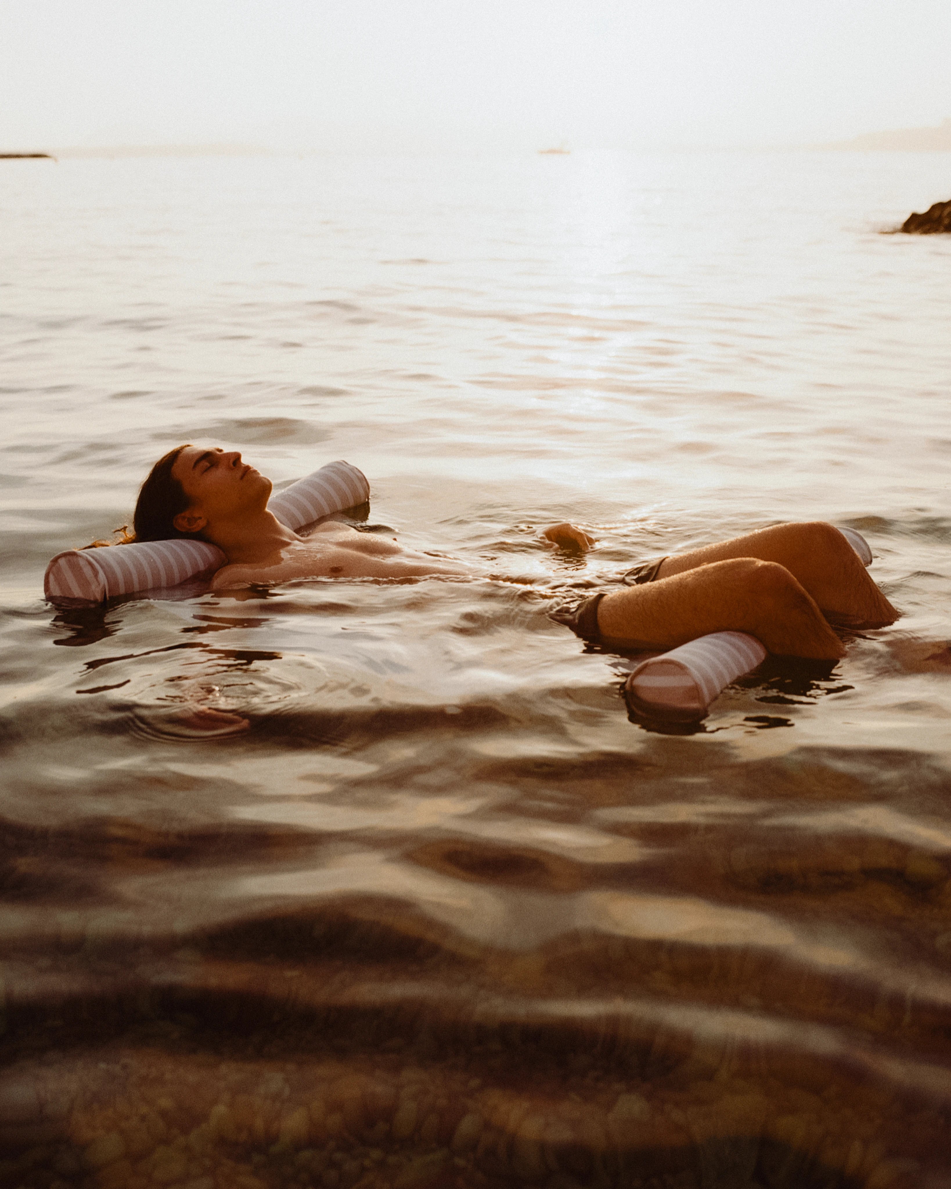 A man floats effortlessly in the sea at golden hour, supported by a striped luxury floating hammock from Oliver James, embodying serene, slow luxury in nature’s embrace.
